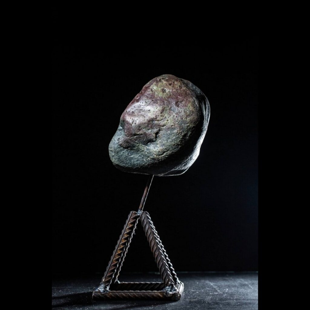 A large, rough stone with a dark, uneven surface is displayed on a metal stand against a black background. Captured by photographer Elena Popkova, the triangular stand made of twisted metal supports the rock at an angle, highlighting its texture and natural shape under subtle lighting.