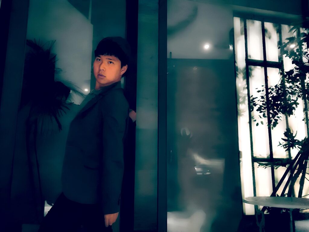 A young man in a dark suit leans against a glass wall in a dimly lit room. The background features frosted glass with backlighting, creating a soft glow. Silhouetted plants add a touch of nature, and the overall atmosphere is modern and moody.