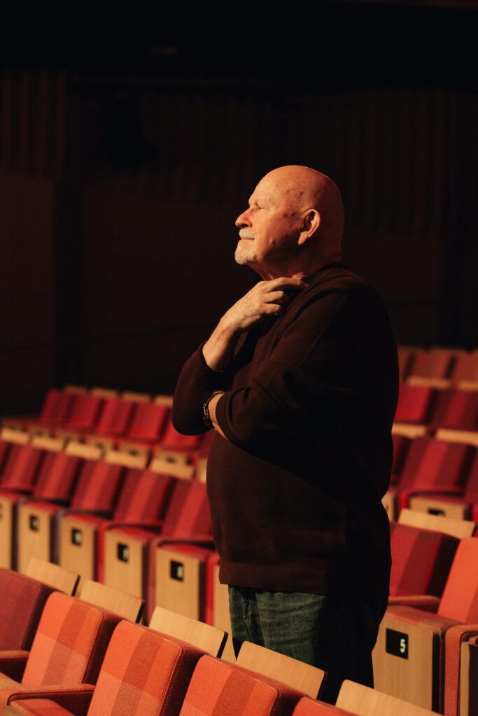 Pēteris Vasks standing among red auditorium seats, looking to the side.