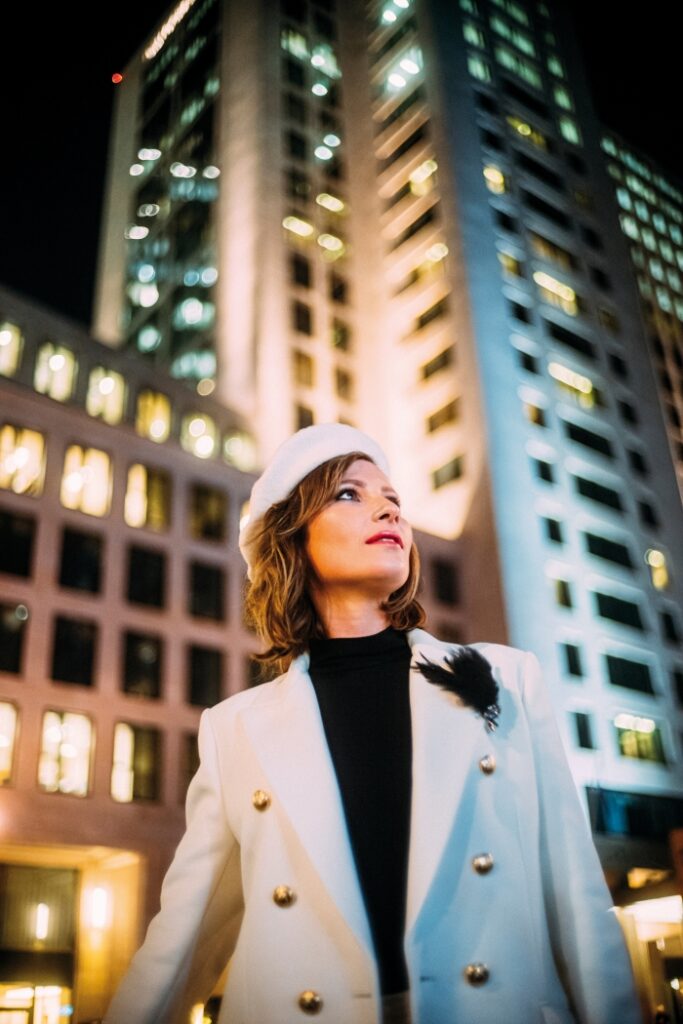 Georgian-born German violinist Lisa Batiashvili in a white jacket and beret against illuminated city buildings, photographed by André Josselin.