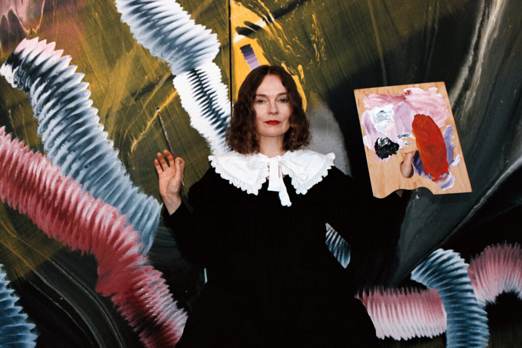 Portrait of Estonian artist Merike Estna holding a painted palette before an abstract mural, photographed by Marta Vaarik